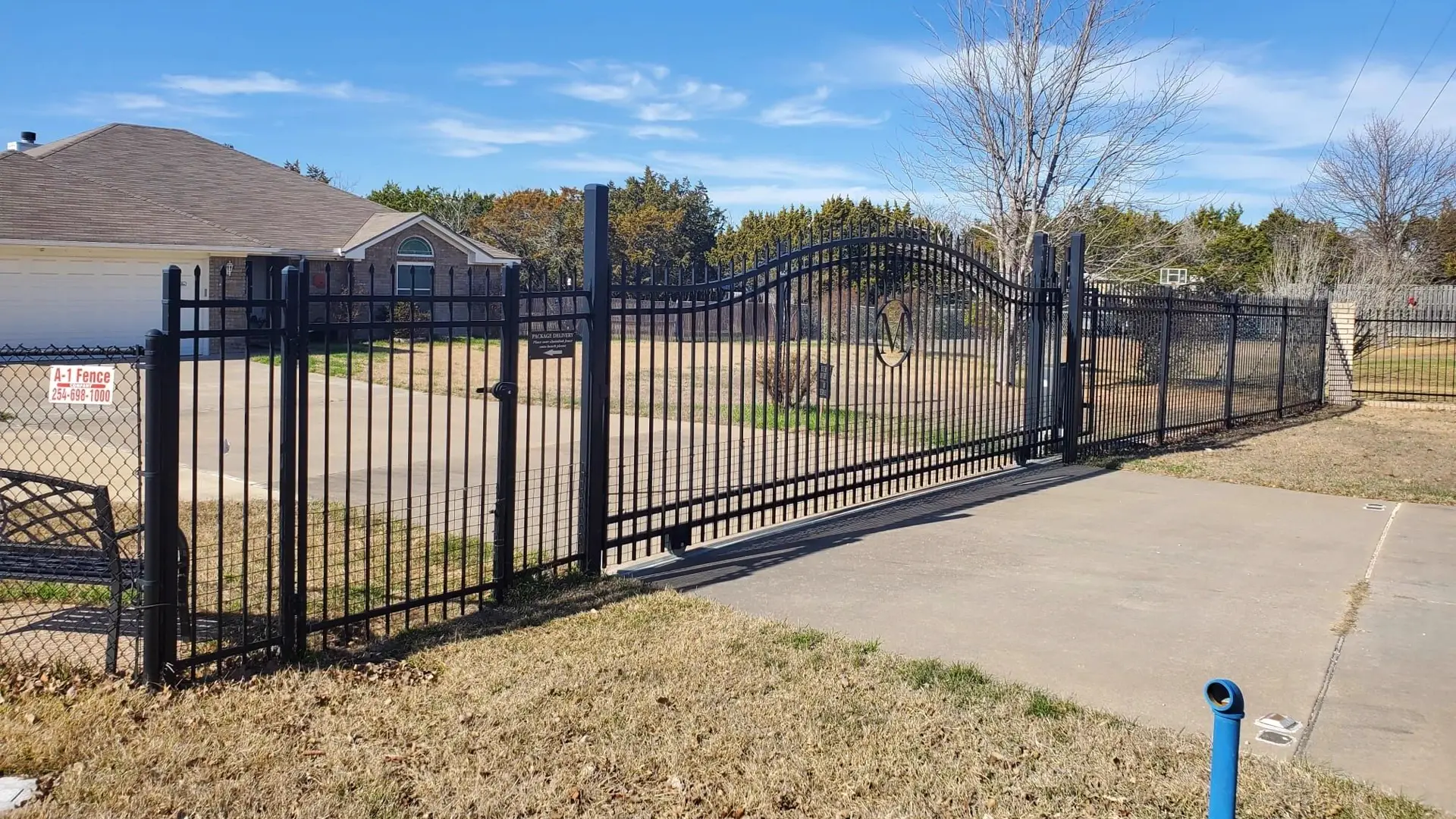 Black ornamental sliding gate with an arched top, set in a black iron fence beside a brick house