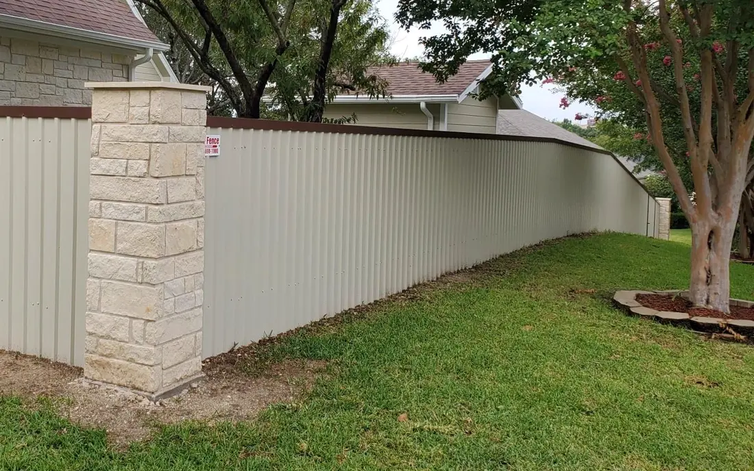 Cream metal privacy fence running along a lawn beside a stone column and trees